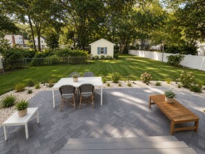 A patio with a table and chairs, surrounded by a herringbone-patterned floor, a wooden bench, and a small garden area with plants.