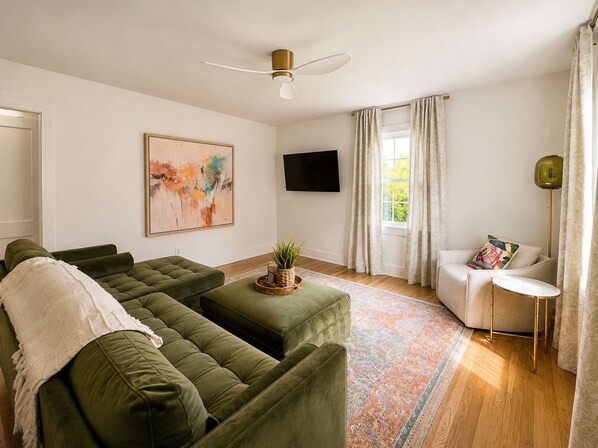 A living room with a green sofa set, a flat-screen TV, a ceiling fan, a window with curtains, and a floor lamp.