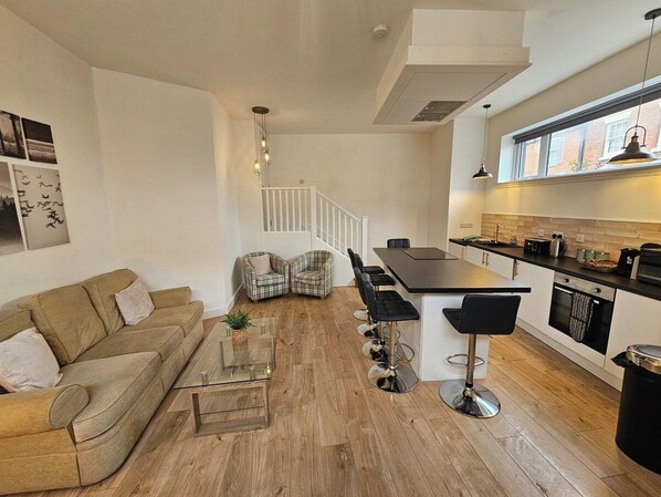 A modern living room with a sofa, chairs, a coffee table, and a kitchen area with a bar and seating. - Kings Central 3 Bedroom - Free Parking & wifi (Southport)