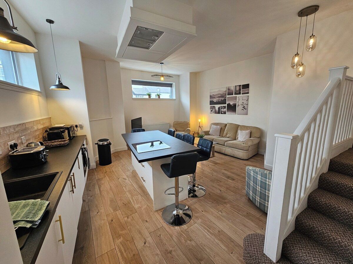 A modern kitchen with a central island, a dining area with bar stools, a sofa, and a staircase leading to another level.