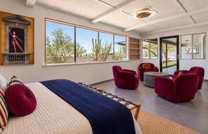 Ein modernes Schlafzimmer mit einem großen Bett, einem Fenster mit Blick auf Kakteen und roten Sitzmöbeln.