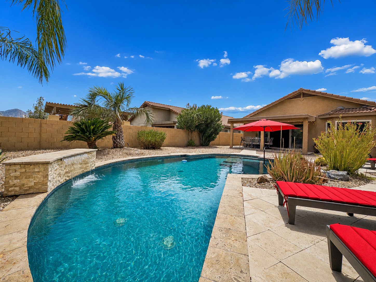 A swimming pool with a curved edge, surrounded by a patio area with red lounge chairs and a red umbrella.