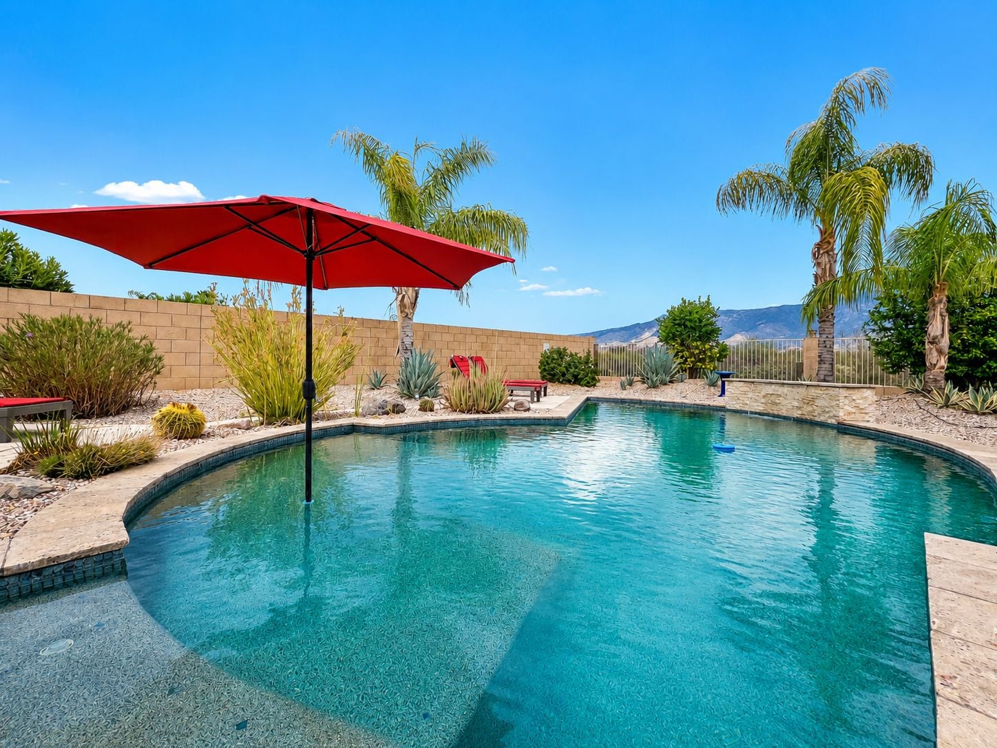 A swimming pool with a red umbrella, surrounded by palm trees and desert plants.