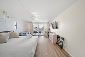 A modern hotel room with a large bed, a desk, a flat-screen TV, and a view of the city. - 36FL New Cozy Apartment Great View Central Waikiki (Honolulu)