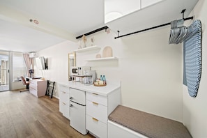A modern hotel room with a kitchenette, a vanity with a mirror, a seating area with a bench, and a view of the city. - 36FL New Cozy Apartment Great View Central Waikiki (Honolulu)