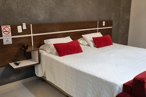 A hotel room with a bed, two red pillows, a white bedspread, a wooden headboard, a shelf with a phone, and a 'No Smoking' sign. - HOTEL VIZON I  (Vilhena)
