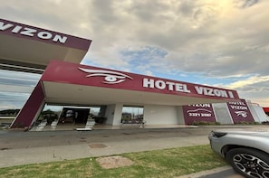 Hotel Vizon, com um logotipo de olho bem visível, um número de contato e área de estacionamento em frente.