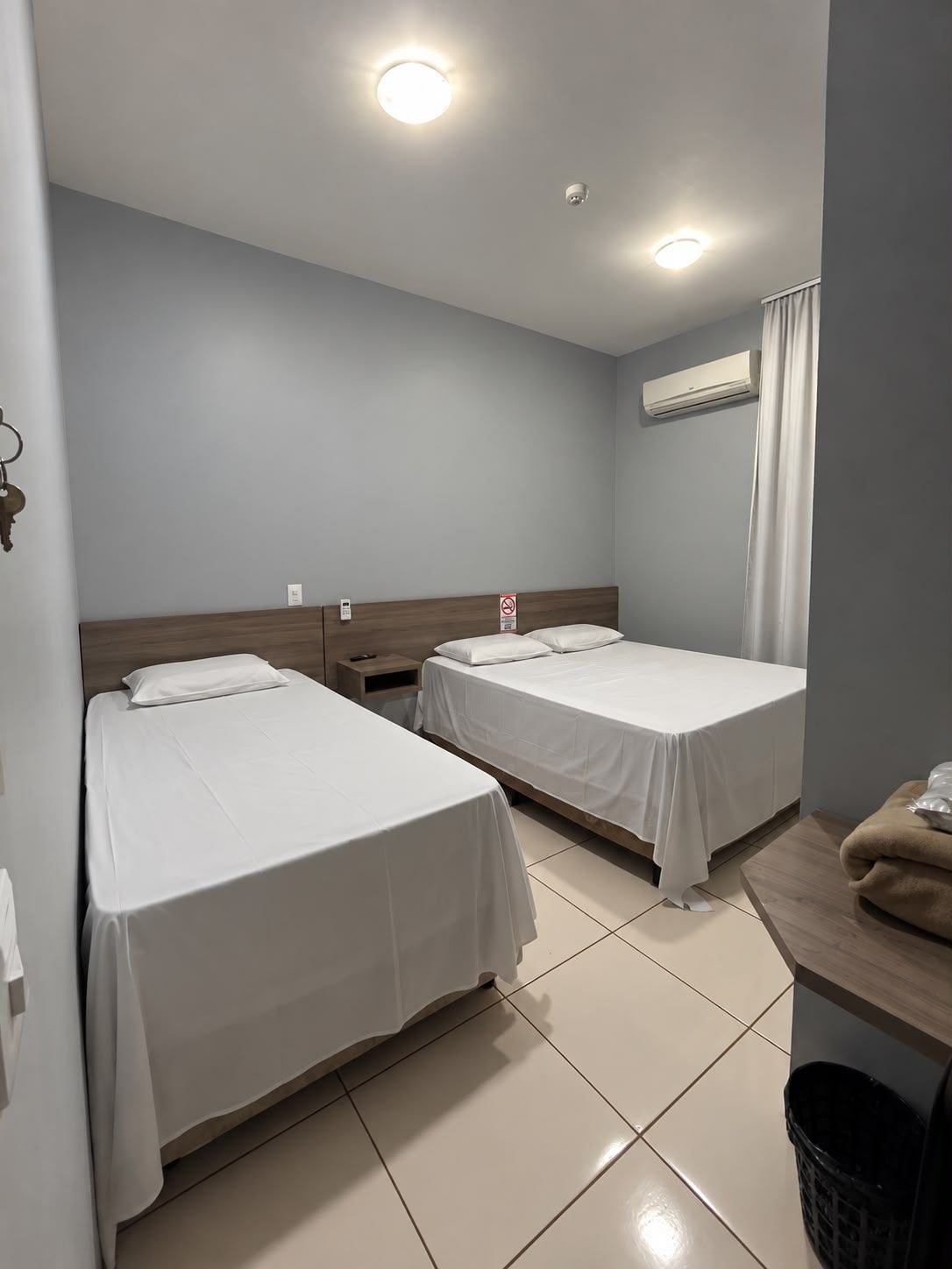 Een hotelkamer met twee enkele bedden, een nachtkastje, een airconditioner, een beveiligingscamera aan de muur en een gordijn.