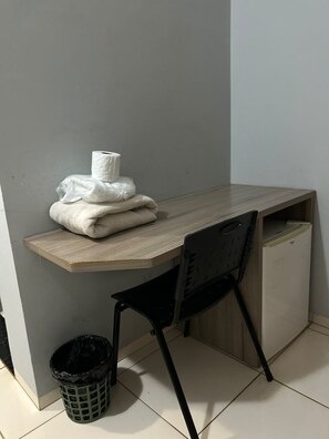 A corner room with a wooden desk, a black chair, a trash bin, and a wall-mounted shelf with a roll of toilet paper.