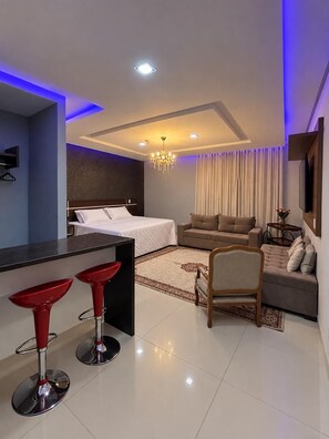 A modern hotel room with a bed, a sofa, a dining area with red bar stools, a chandelier, and a large window with curtains.