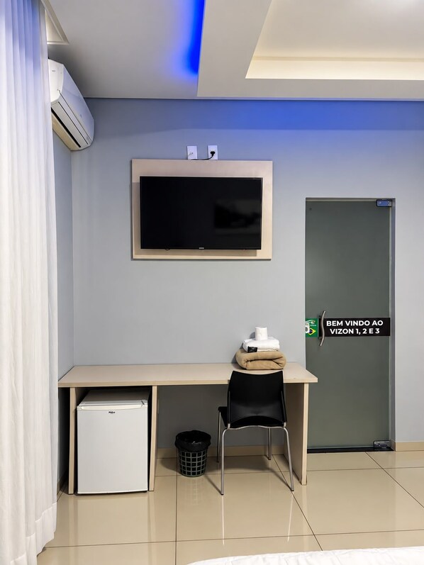 A hotel room with a bed, a desk with a chair, a television, a mini-fridge, a trash bin, and a door with a sign.