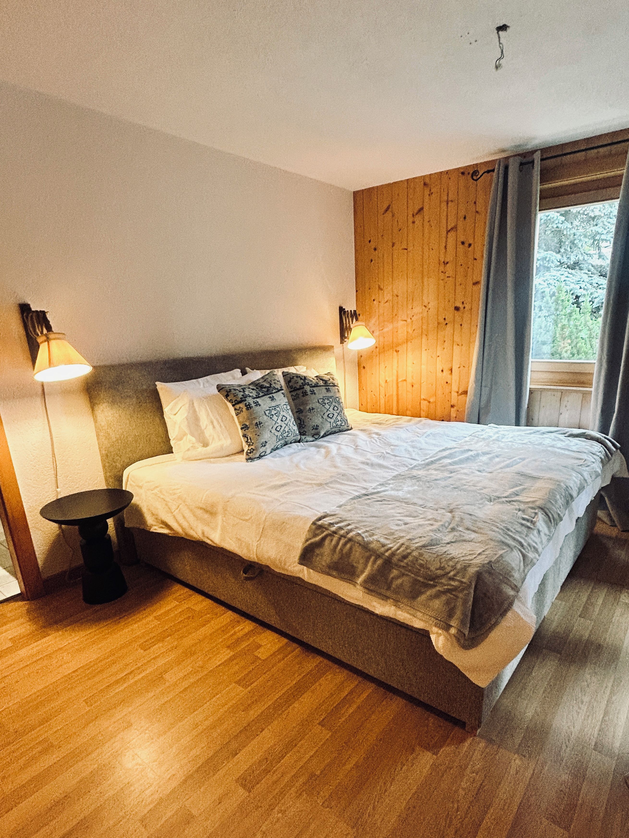 A bedroom with a wooden headboard, a bed with patterned pillows, two bedside lamps, and a window with curtains.