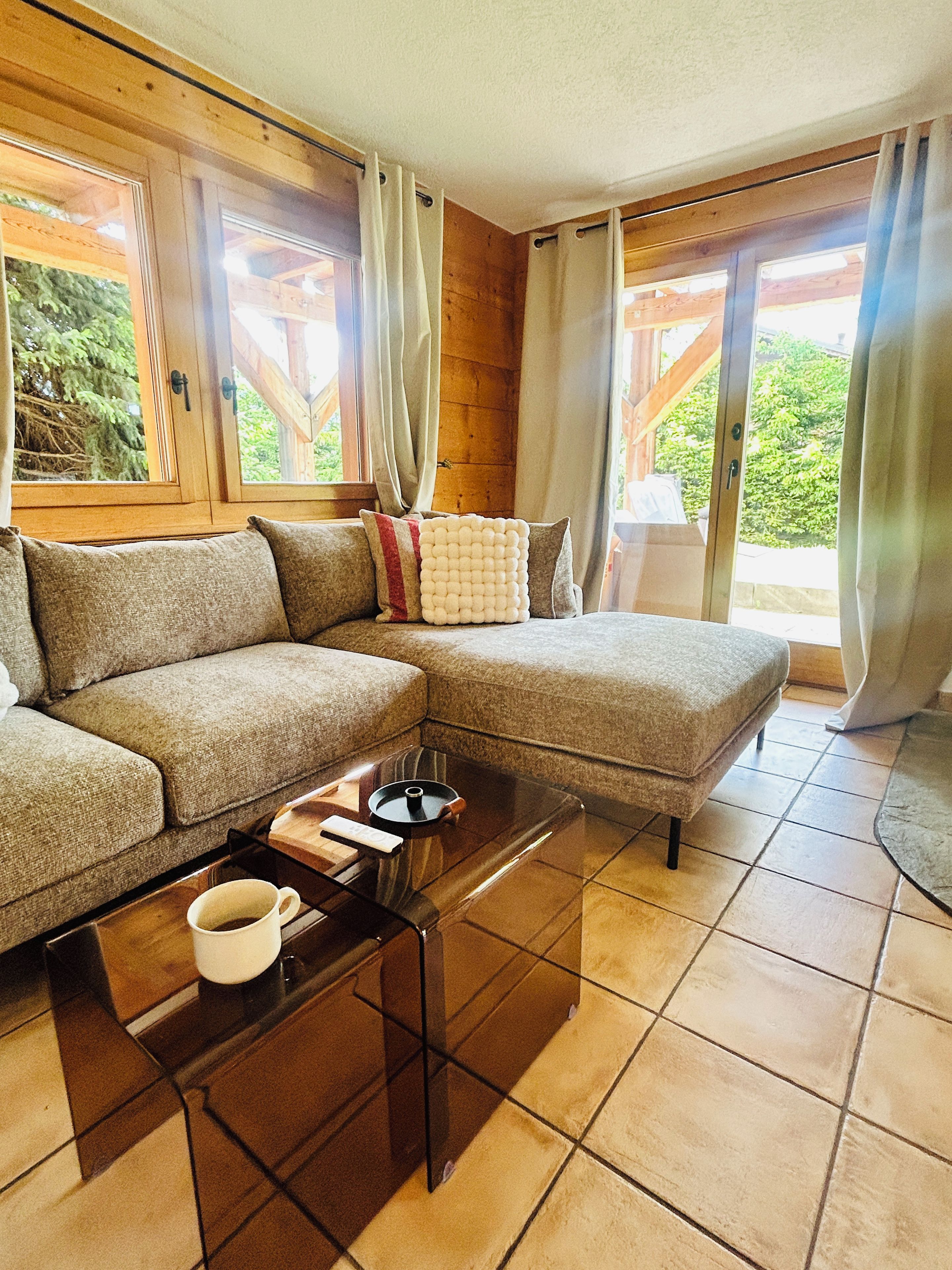 A modern living room with a sectional sofa, a glass coffee table, and a view of the outdoors through large windows.
