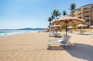 Ein Strand mit Liegestühlen und Sonnenschirmen, einem klaren Himmel und einem Gebäude im Hintergrund.