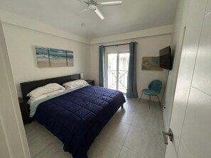 A bedroom with a bed, a ceiling fan, a window with curtains, a chair, and a painting on the wall. - Sunset Cove Condo 314 Beach View Bliss-2-Bedroom (George Town)