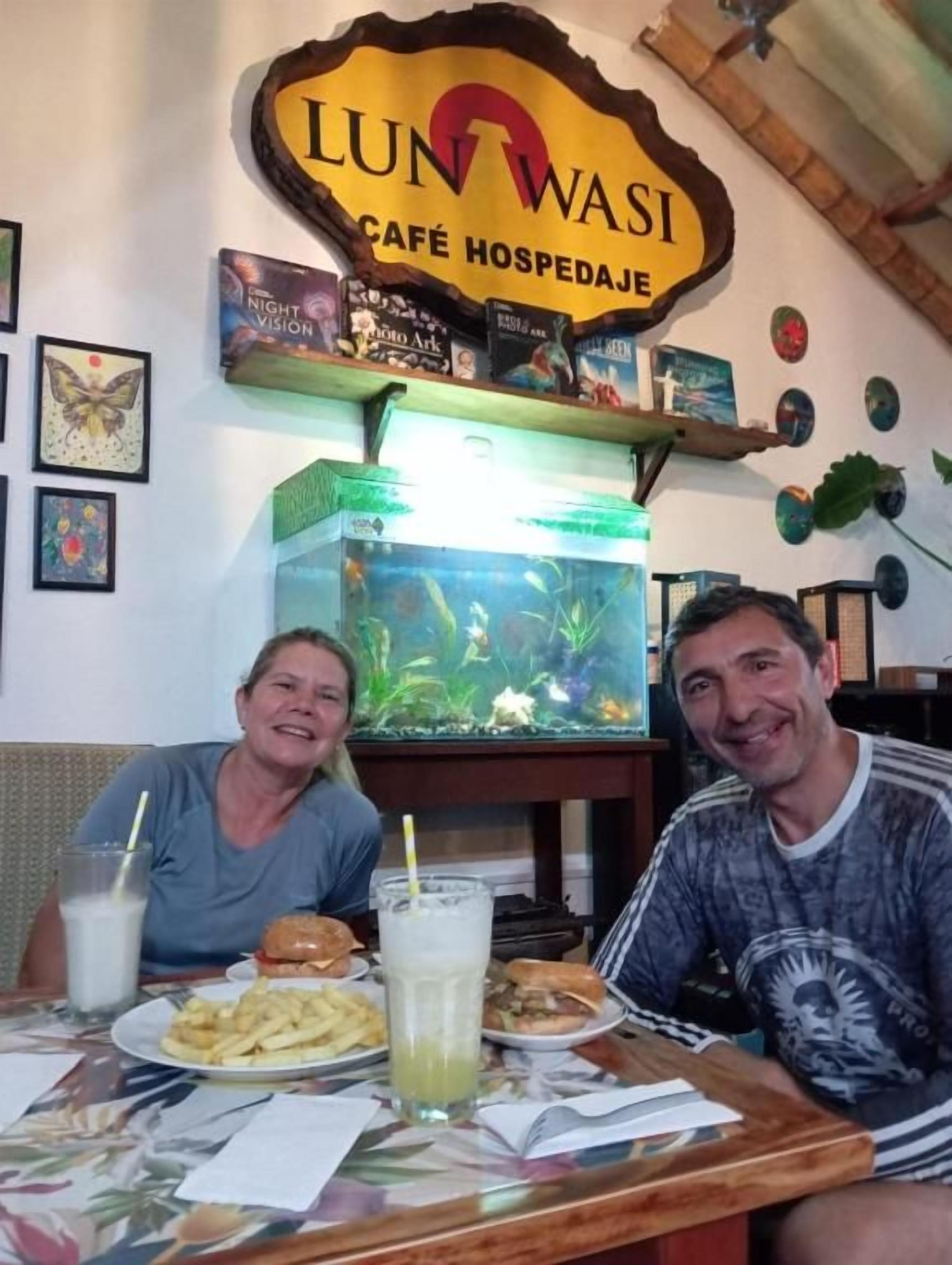 A café with a fish tank, a sign reading 'LUN WASI CAFÉ HOSPEDAJE', and a table with food and drinks.