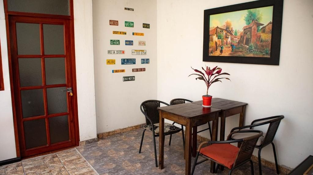 A room with a wooden door, a tiled floor, a table with chairs, a potted plant, and a framed painting on the wall.