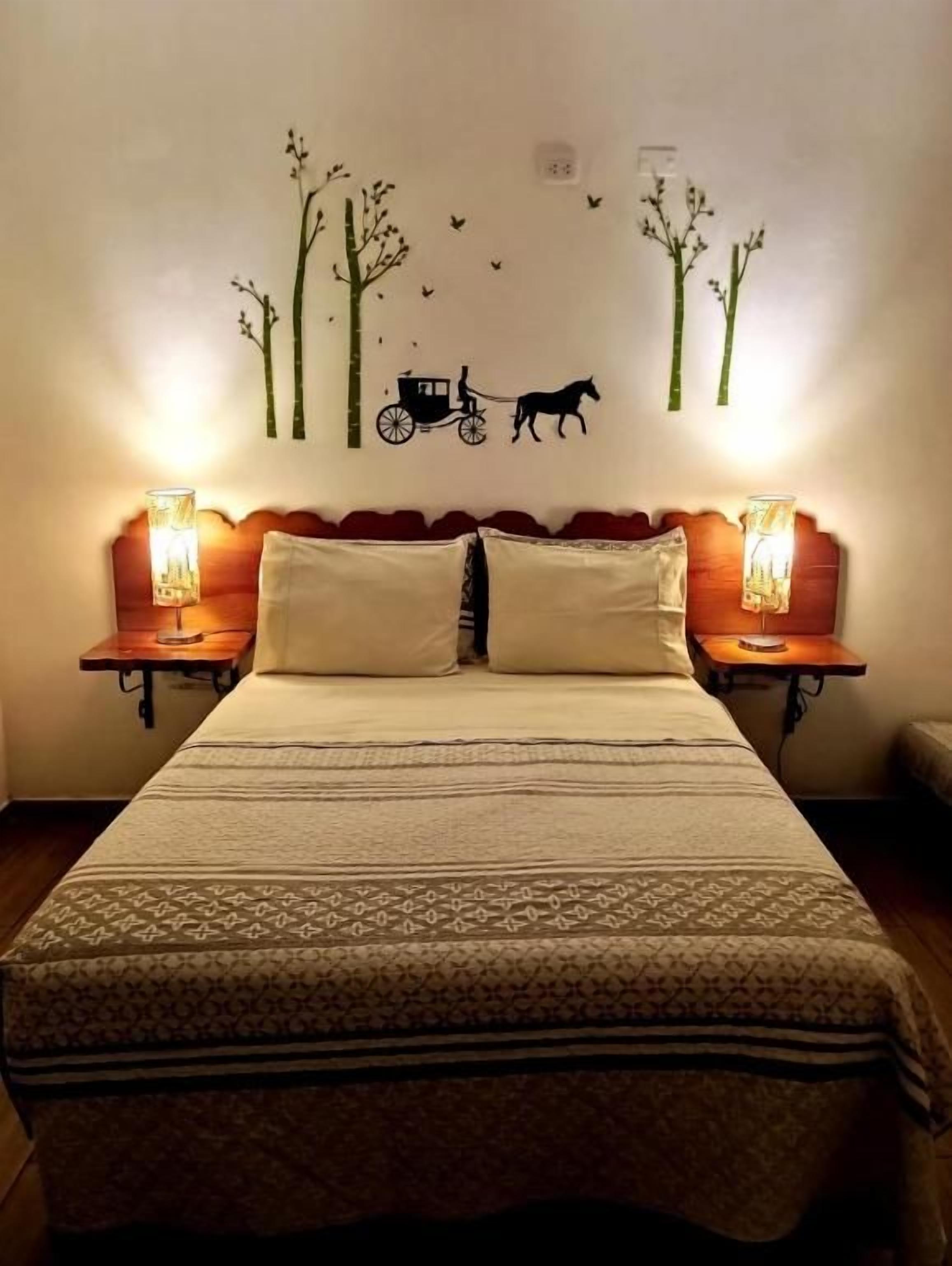 A bed with a patterned bedspread, two bedside lamps, and a wall decal of trees and a horse-drawn carriage.