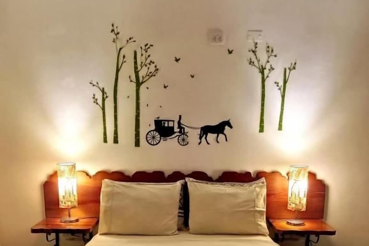 A bed with a patterned bedspread, two bedside lamps, and a wall decal of trees and a horse-drawn carriage.