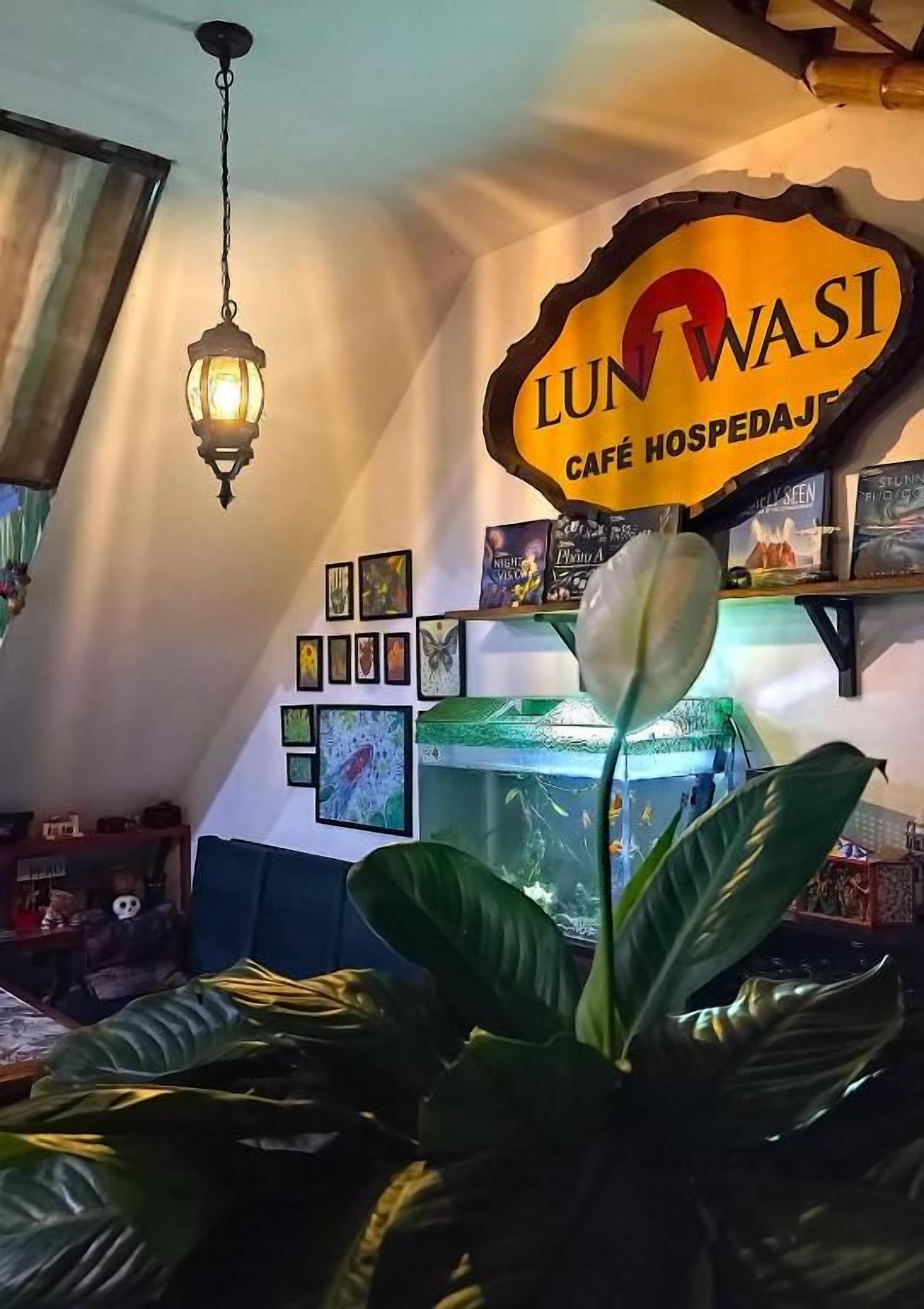 A café interior with a sign reading 'LUN WASI CAFÉ HOSPEDAJER,' a hanging light fixture, and a fish tank.