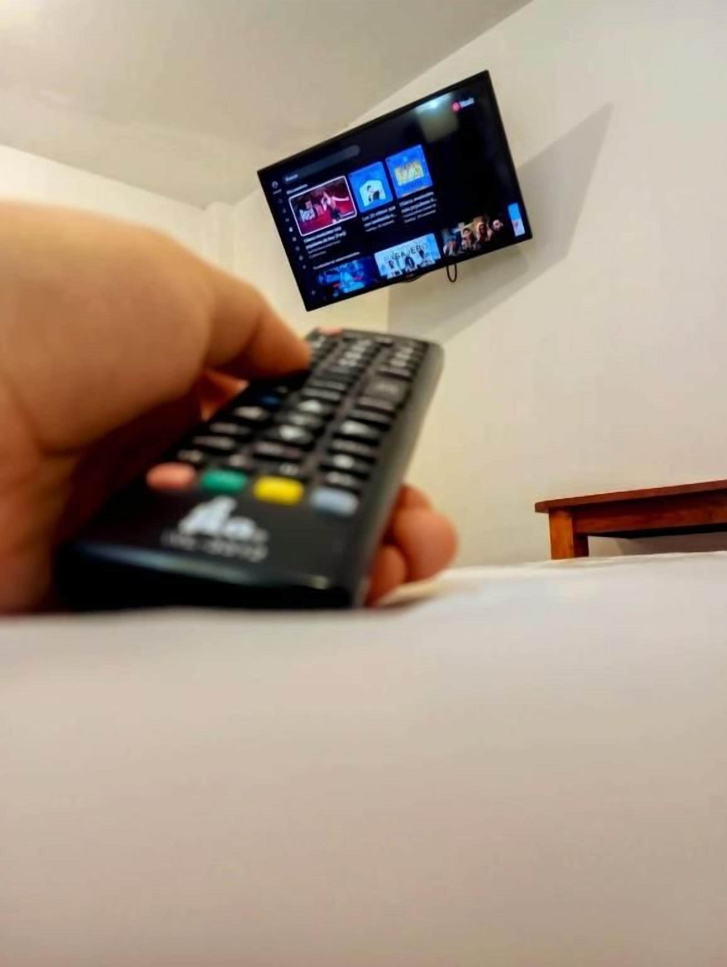 A hand holding a remote control in front of a television mounted on a wall.
