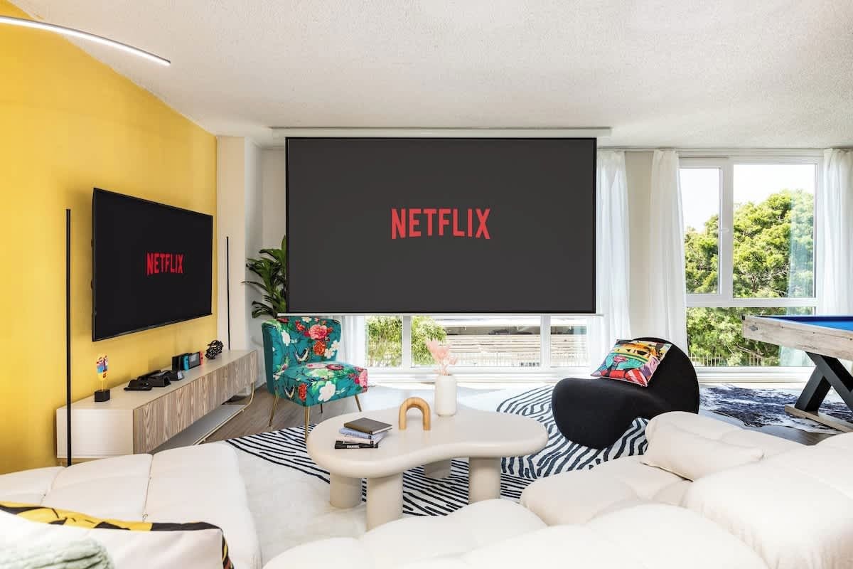 A modern living room with a large flat-screen TV displaying 'NETFLIX,' a sofa, a coffee table, and a window with a view of greenery.