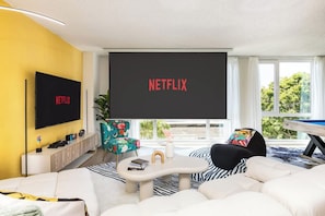 A modern living room with a large flat-screen TV displaying 'NETFLIX,' a sofa, a coffee table, and a window with a view of greenery. - Ocean|Romantic Bath|Cinema|Pool|Arcade|Grill (Marina del Rey)
