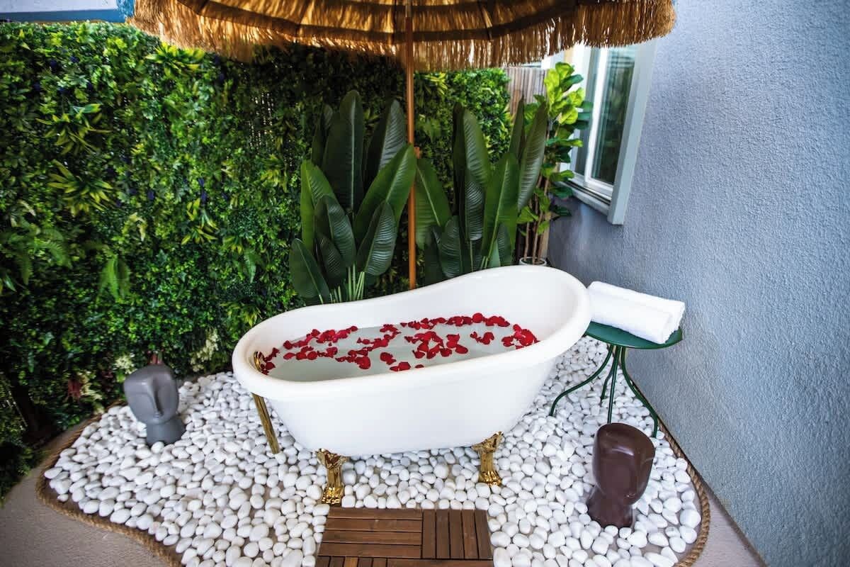 Une baignoire remplie de pétales de roses rouges, entourée de petits galets blancs, avec un tapis vert et un banc en bois à proximité.
