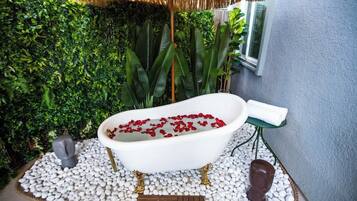 Une baignoire remplie de pétales de roses rouges, entourée de petits galets blancs, avec un tapis vert et un banc en bois à proximité.