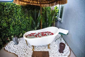 Eine Badewanne, gefüllt mit roten Rosenblättern, umgeben von weißen Kieselsteinen, mit einer grünen Matte und einer hölzernen Bank in der Nähe.