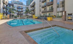 Una piscina con varias pistas, rodeada de sillones de descanso y un edificio de varios pisos con balcones.