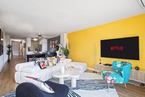 Una sala de estar con un sofá, una mesa de centro y un sillón colorido. Un televisor de pantalla plana montado en la pared muestra 'NETFLIX'.