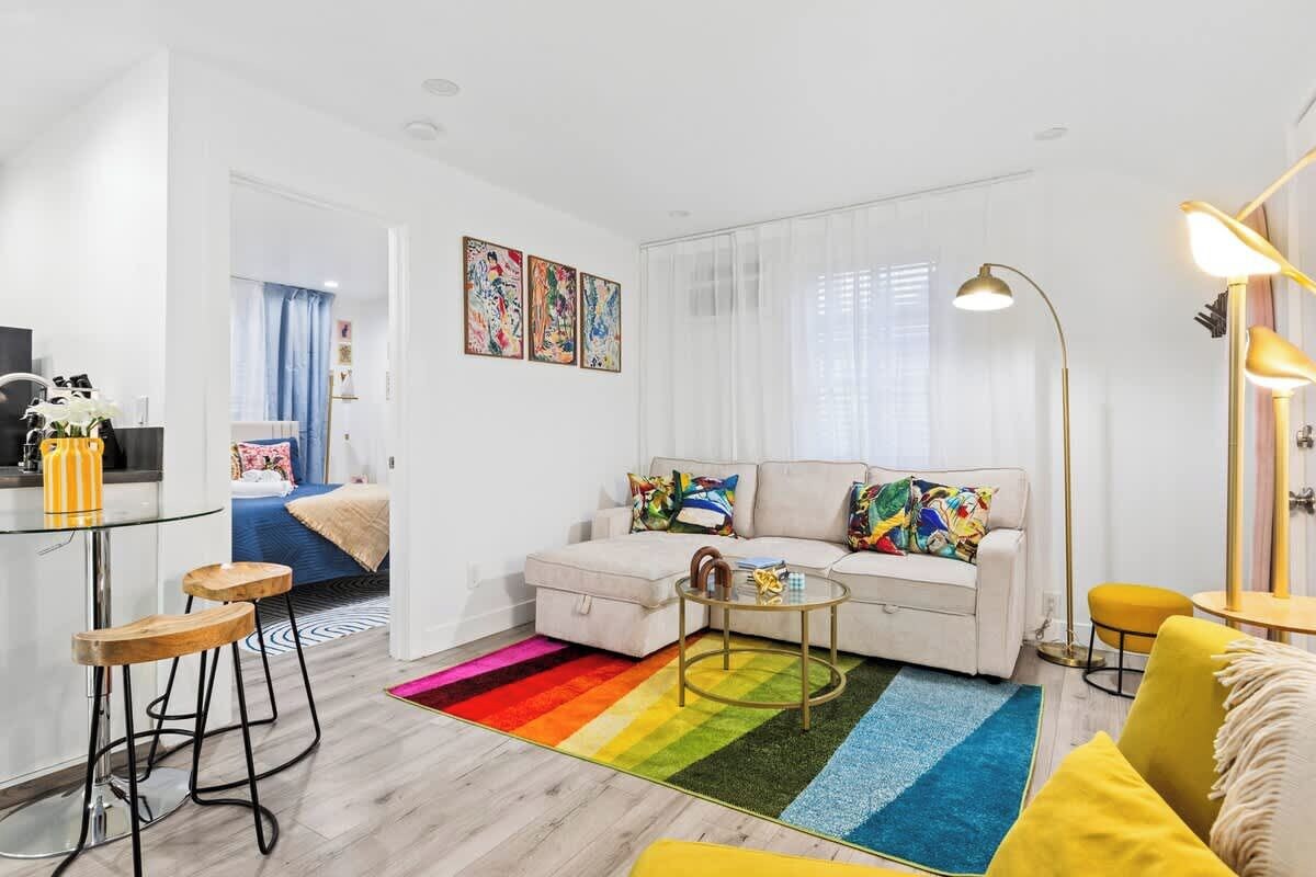 A modern living room with a sofa, coffee table, and a colorful rug. There is a dining area with stools and a kitchen area with a fridge and a sink.