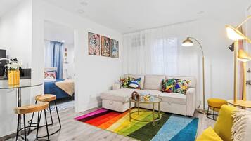 A modern living room with a sofa, coffee table, and a colorful rug. There is a dining area with stools and a kitchen area with a fridge and a sink.