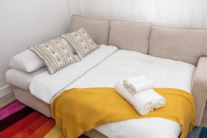 Een bed met wit beddengoed, twee gestreepte kussens, een gele deken en twee opgevouwen handdoeken.