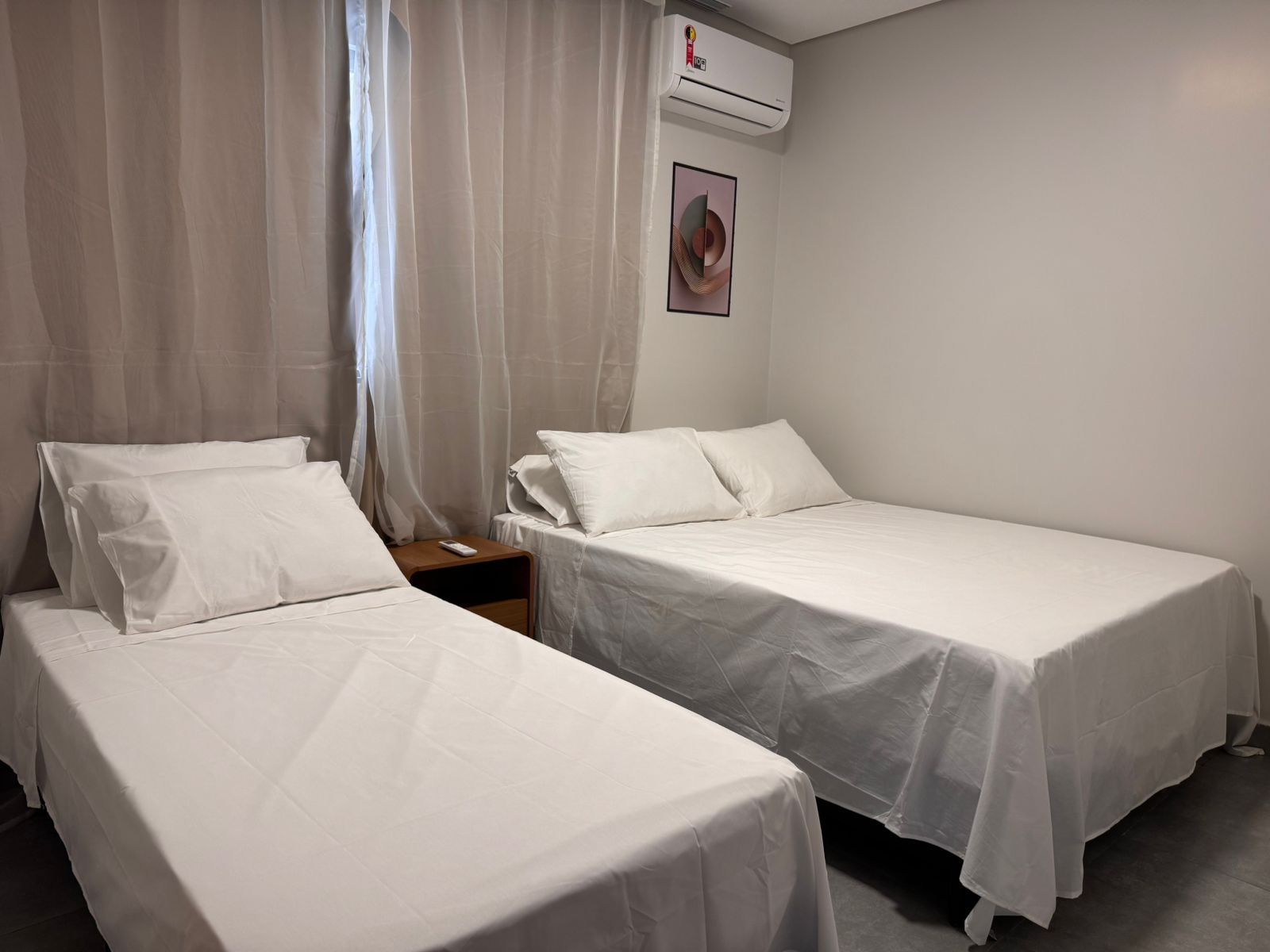 A hotel room with two single beds, a nightstand, a remote control, an air conditioning unit, and a framed picture on the wall.