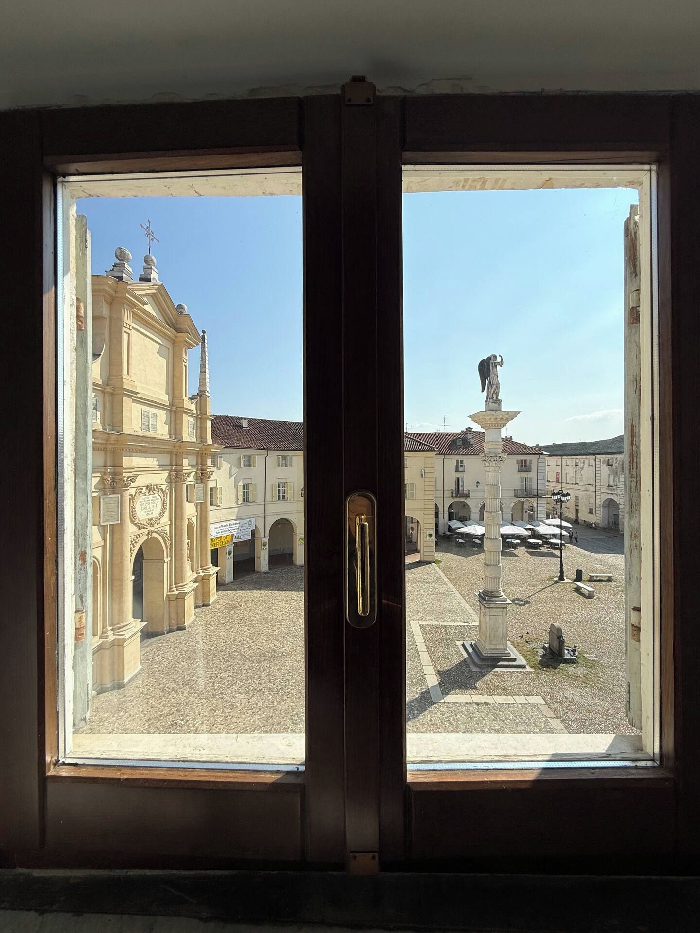 Vista attraverso una finestra con una porta, che mostra una piazza con una fontana, edifici e cielo sereno.