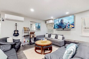 A modern living room with a flat-screen TV, a guitar, a bookshelf, and a round wooden coffee table. - Peace at the River -Private Dock, Games, & FirePit (Bethel Island)