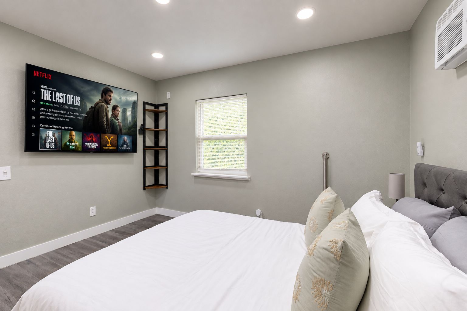 A bedroom with a large bed, a TV mounted on the wall, a window with blinds, and a radiator.