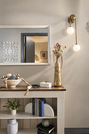 A modern interior with a wooden console table, a decorative mirror, wall-mounted lights, and a bookshelf with books and decorative items. - The Bentley Collection - The Landmark Loft (Dallas)