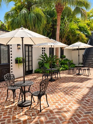 An outdoor patio with brick flooring, black metal chairs and tables, and white umbrellas providing shade.