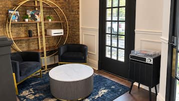 Une pièce avec un mur en briques, une porte noire, une table ronde, deux chaises noires, une bibliothèque et un petit meuble noir.