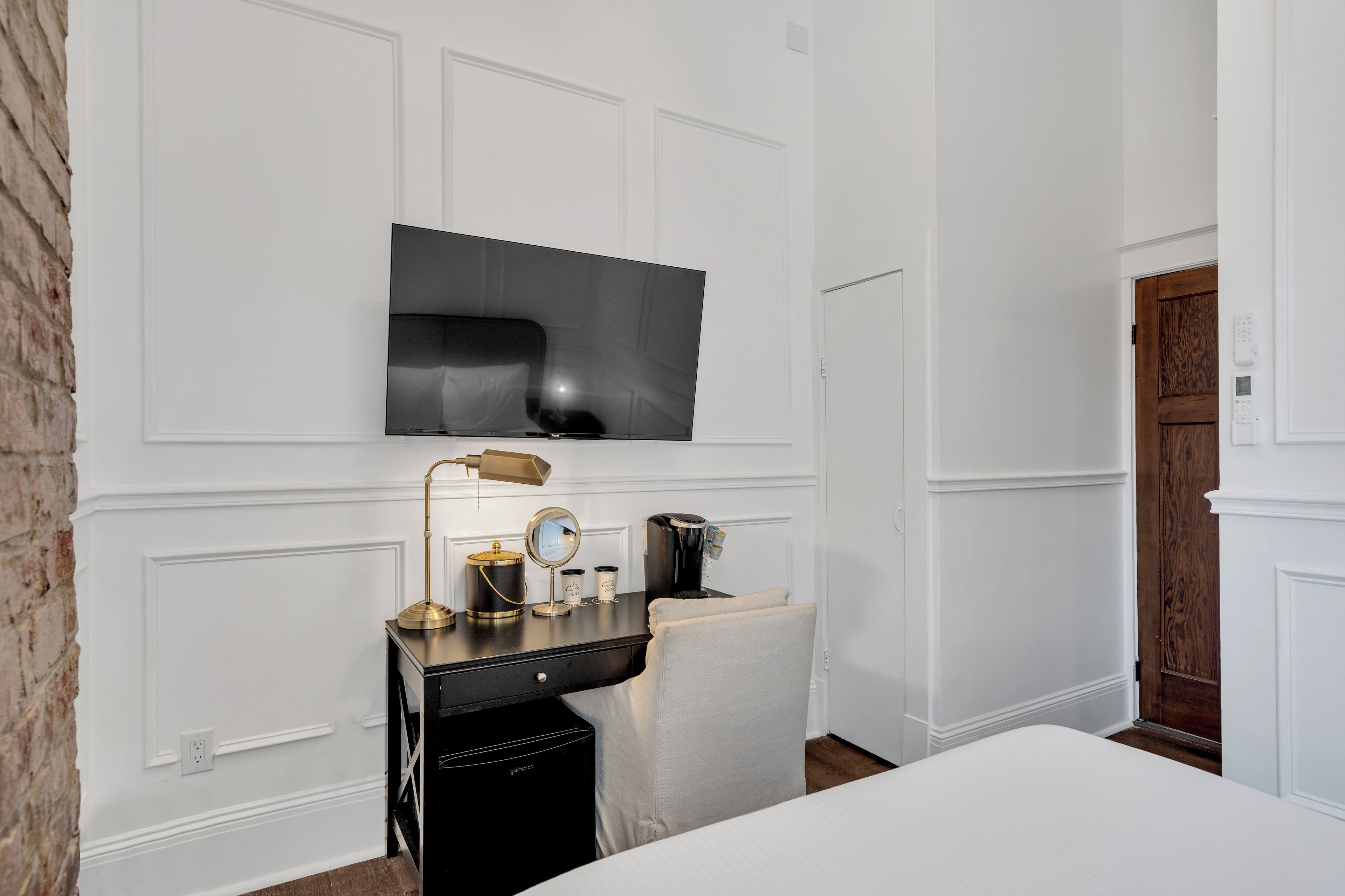A hotel room with a flat-screen TV mounted on the wall, a desk with a lamp, a coffee maker, and a small mirror.