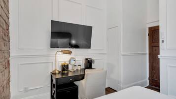 A hotel room with a flat-screen TV mounted on the wall, a desk with a lamp, a coffee maker, and a small mirror.