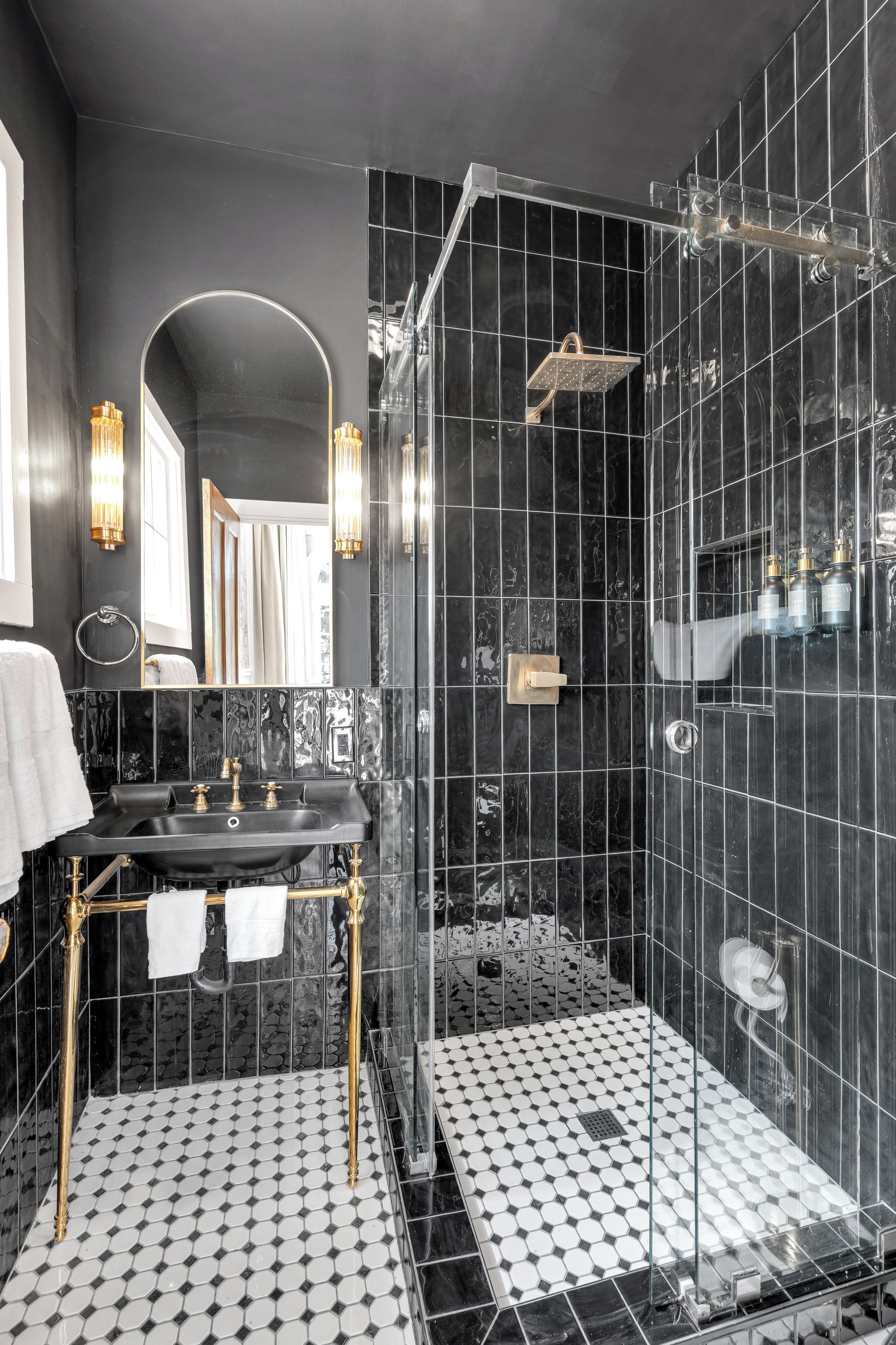 Ein modernes Badezimmer mit schwarz-weiß gefliestem Boden, einem goldenen Waschtisch und einer Duschkabine aus Glas. Über dem Spiegel sind Wandleuchten angebracht.