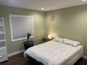 A bedroom with a bed, a bedside table with a lamp, a window with blinds, and a wall clock. - Spacious 4-bedroom house in beautiful Elliot Lake. Close to all trails and town. (Elliot Lake)
