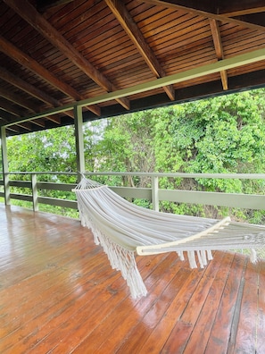 A hammock under a wooden roofed deck with a view of green foliage.