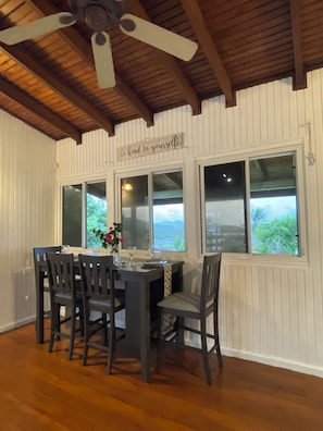 A dining area with a table set for four, wooden chairs, a ceiling fan, and a view through the windows.