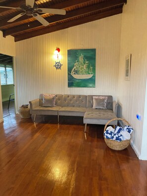 Een woonkamer met een hoekbank, een plafondventilator, een schilderij van een schip aan de muur en een rieten mand.