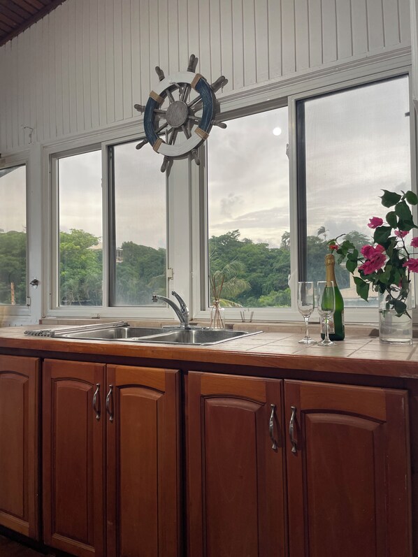 A kitchen with a large window, wooden cabinets, a stainless steel sink, and a ship's wheel decoration.